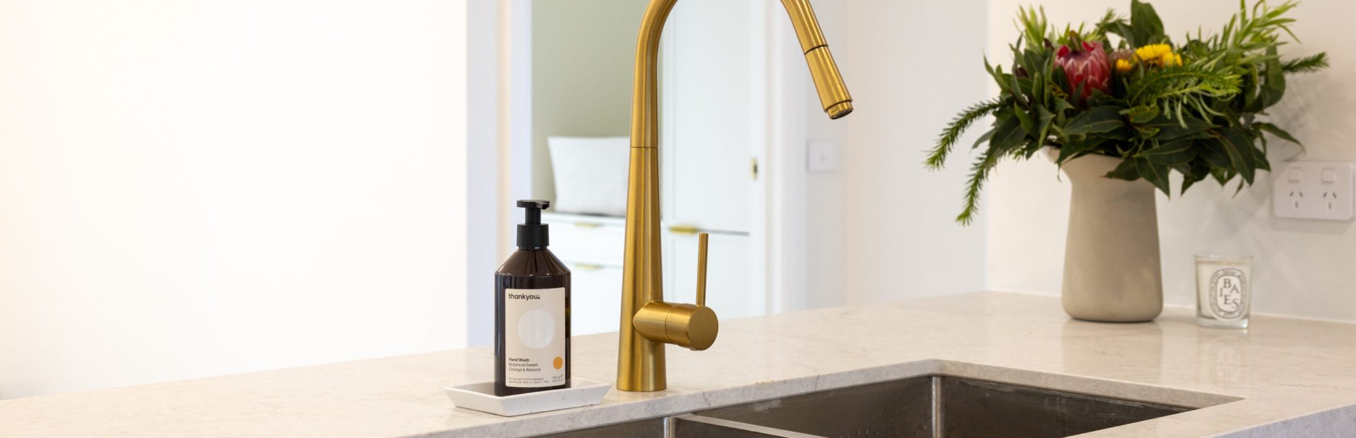 White kitchen cabinetry with metal sink and brass tapware with flowers and hand soap on stone benchtop