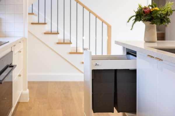 Double kitchen bins in grey pulled out with white hampton kitchen cabinet door next to timber staircase with oak timber flooring
