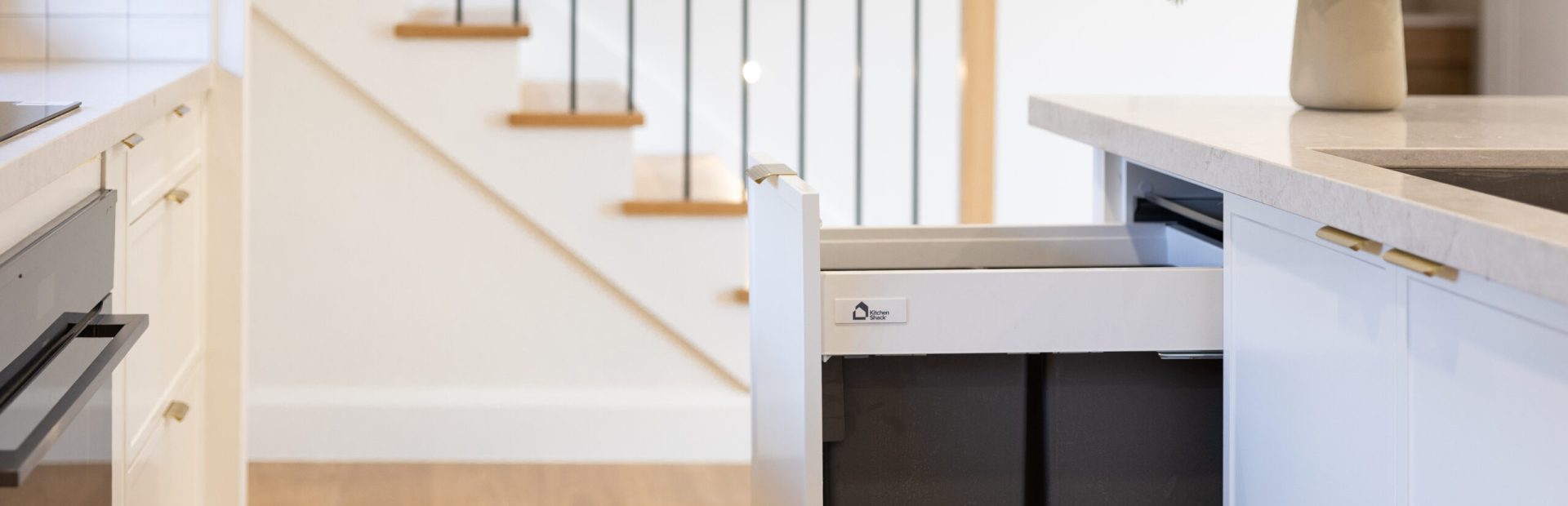Double kitchen bins in grey pulled out with white hampton kitchen cabinet door next to timber staircase with oak timber flooring