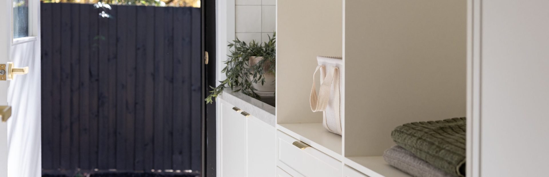 Laundry displaying white kitchen hampton cabinetry with brass handles and open shelves displaying green towels and white basket flowing to open door to external deck