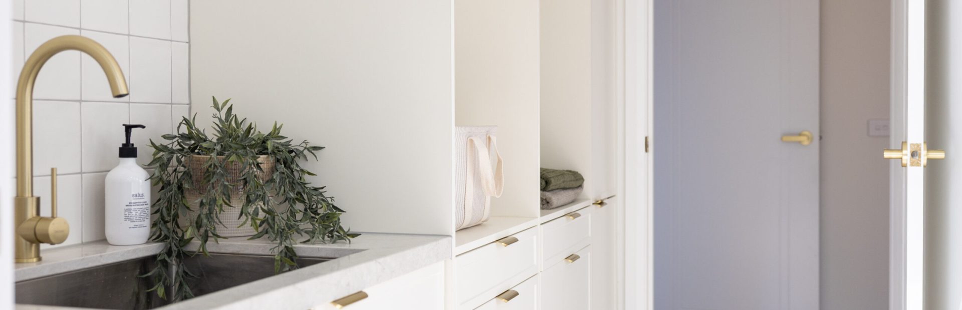 White hampton style laundry cabinetry with brass handles in this galley laundry with a plant on stone benchtop.