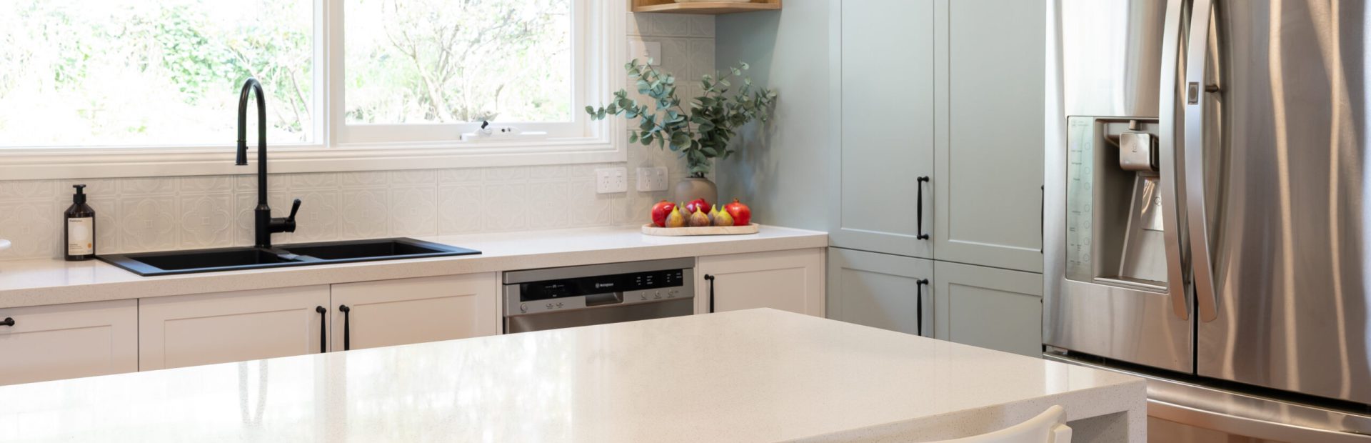 Green, timber and white kitchen with a large island bench with white benchstools and black sink and tap looking out the kitchen window
