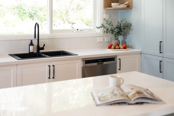 Galley style kitchen with white kitchen cabinetry and black handles with island bench with coffee cup and recipe book open