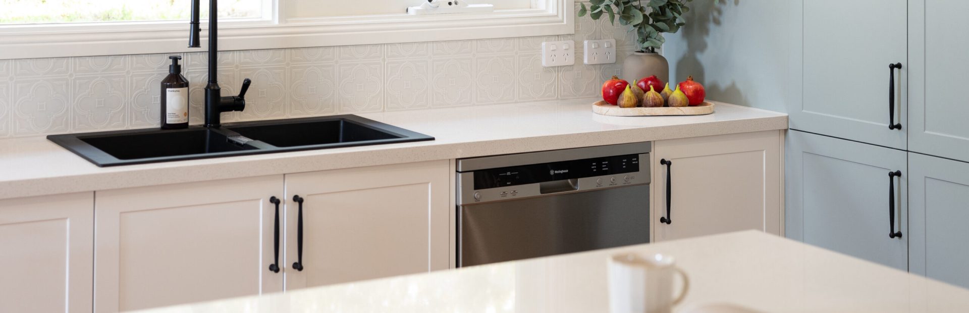 Galley style kitchen with white kitchen cabinetry and black handles with island bench with coffee cup and recipe book open