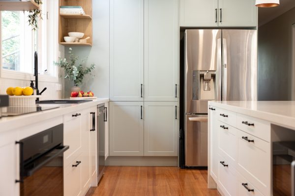 White and green galley kitchen with black handles and end pantry next to an open shelf displaying cookbooks and fruit and folliage on bench