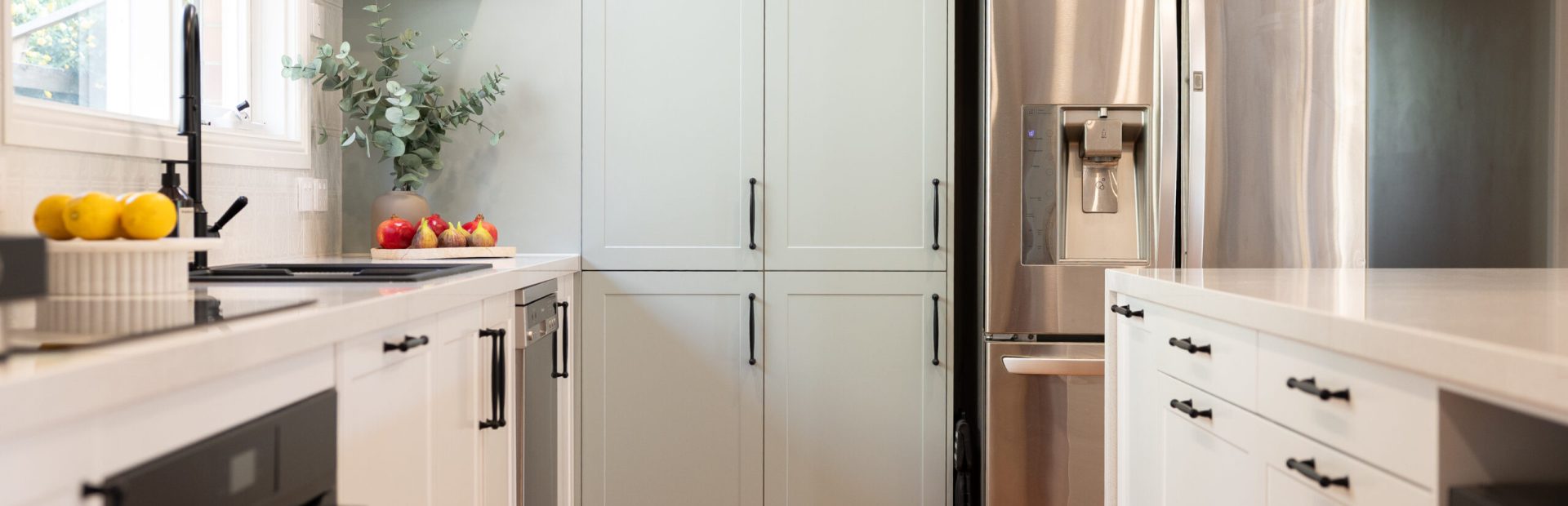 White and green galley kitchen with black handles and end pantry next to an open shelf displaying cookbooks and fruit and folliage on bench