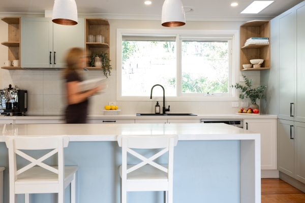 Lady holding cookbook walking through galley style kitchen with timber open shelves and green island bench with white benchstools