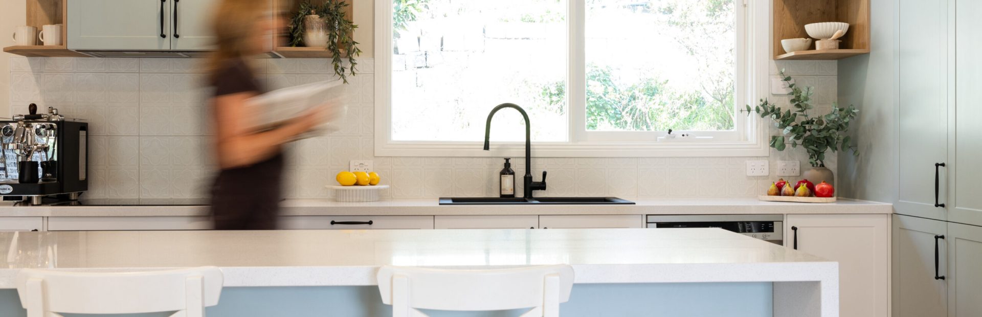 Lady holding cookbook walking through galley style kitchen with timber open shelves and green island bench with white benchstools