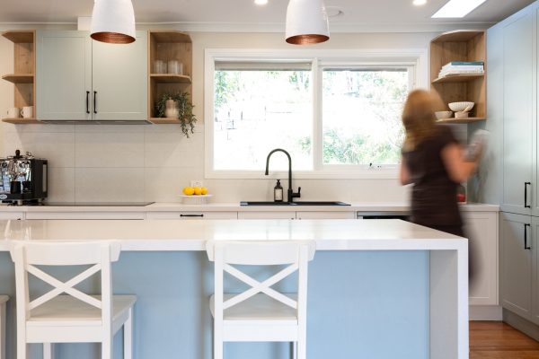 Lady putting cookbook on timber shelf next to a kitchen window displaying black sink and tap and timber open shelf next to an island bench with white benchstools