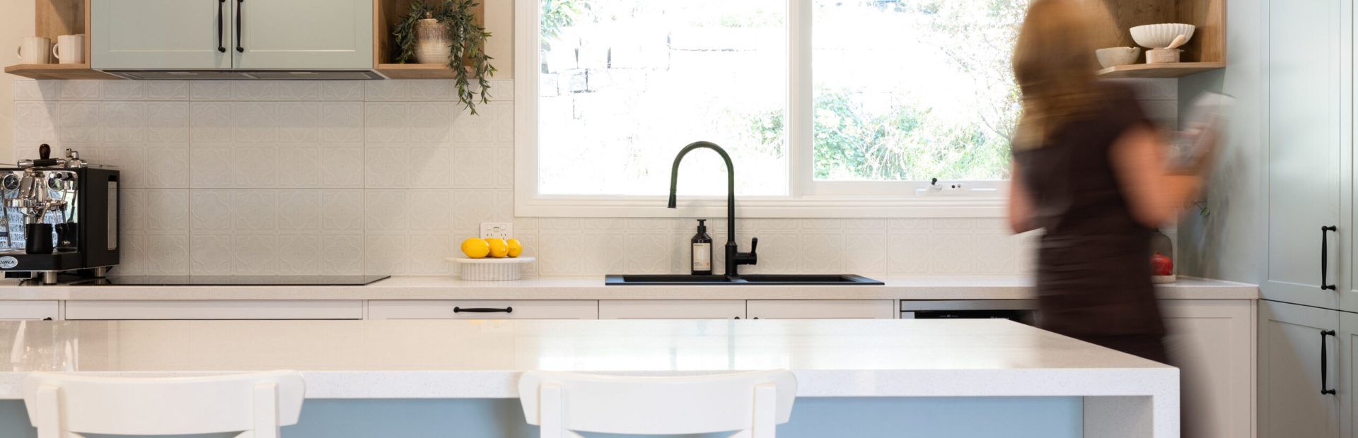 Lady putting cookbook on timber shelf next to a kitchen window displaying black sink and tap and timber open shelf next to an island bench with white benchstools