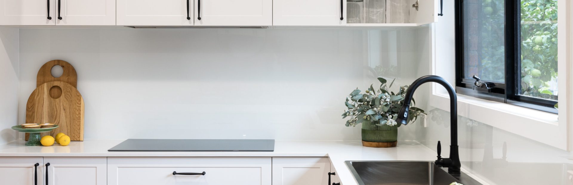 White shaker style kitchen with black handles and black sink and tap with black handles and plant on stone benchtops