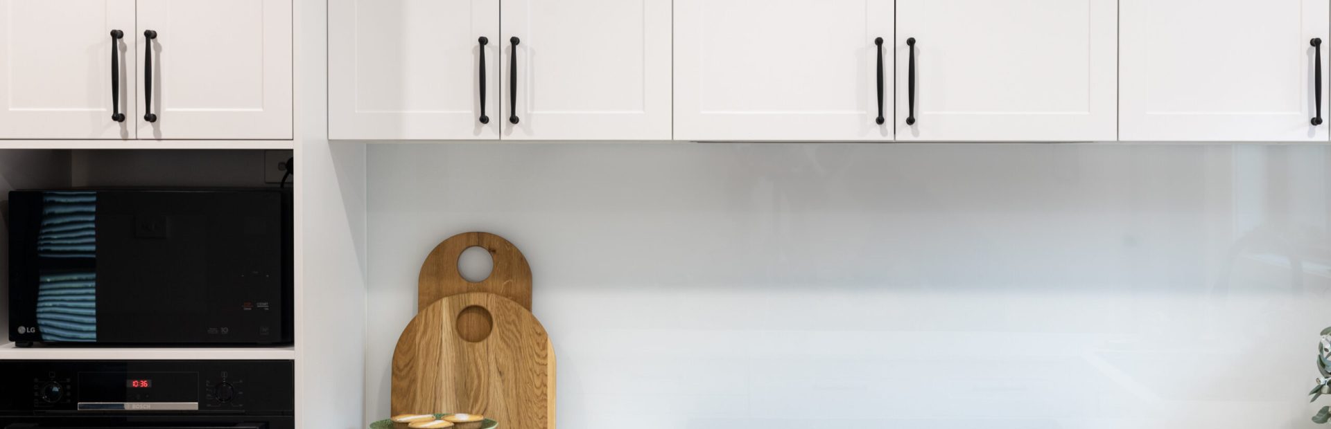 White shaker style kitchen cabinetry with black induction cooktop and timber chopping boards with green stand holding lemon tarts and lemons on stone benchtops