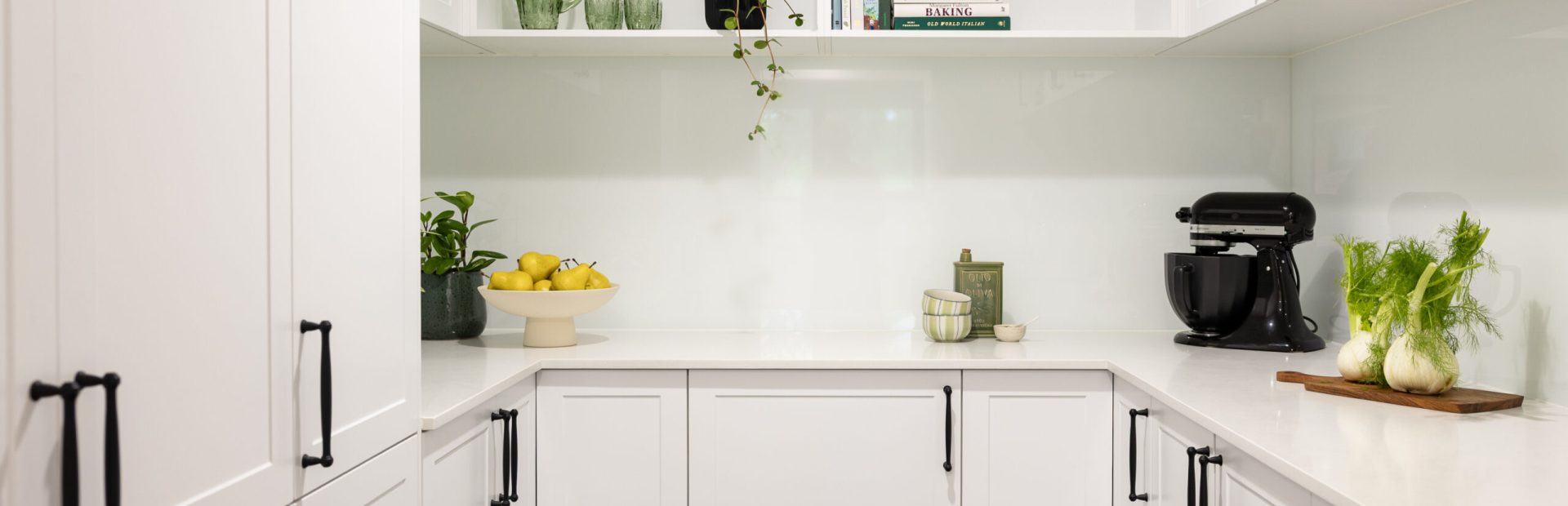 U shaped butlers pantry with white shaker style cabinetry with black handles and open shelves displaying green glasses, plant and styling items on stone benchtops