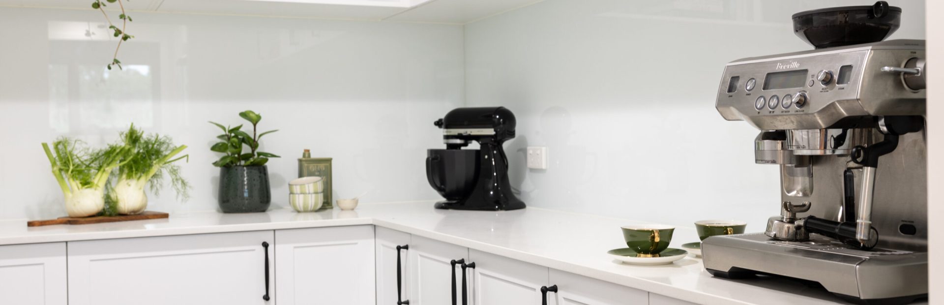 Butlers pantry displaying white shaker style kitchen cabinetry with black decorative handles and black mixmaster, potplant and cookbooks with plants in open white shelving