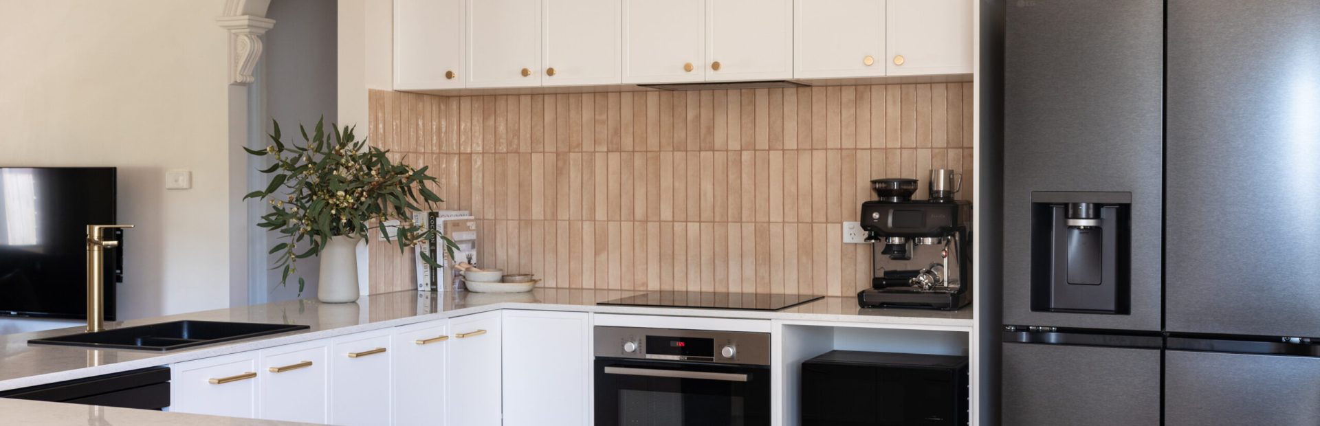 U shape kitchen with white hampton style kitchen cabinetry with brass handles, stone benchtops and black appliances against a pink tiled splashback