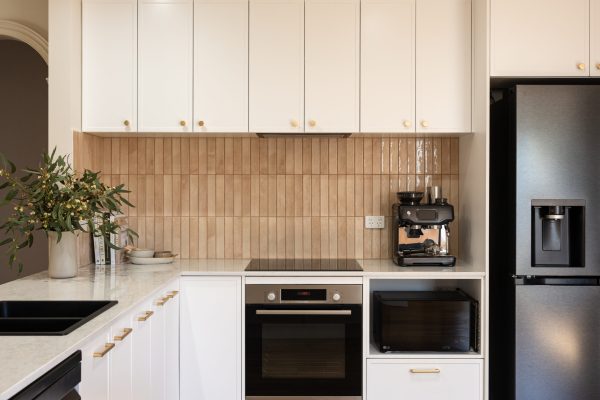 U shape kitchen displaying hampton style white kitchen cabinetry with brass handles and pink tiled splashback with coffee machine on stone benchtops and black oven and microwave