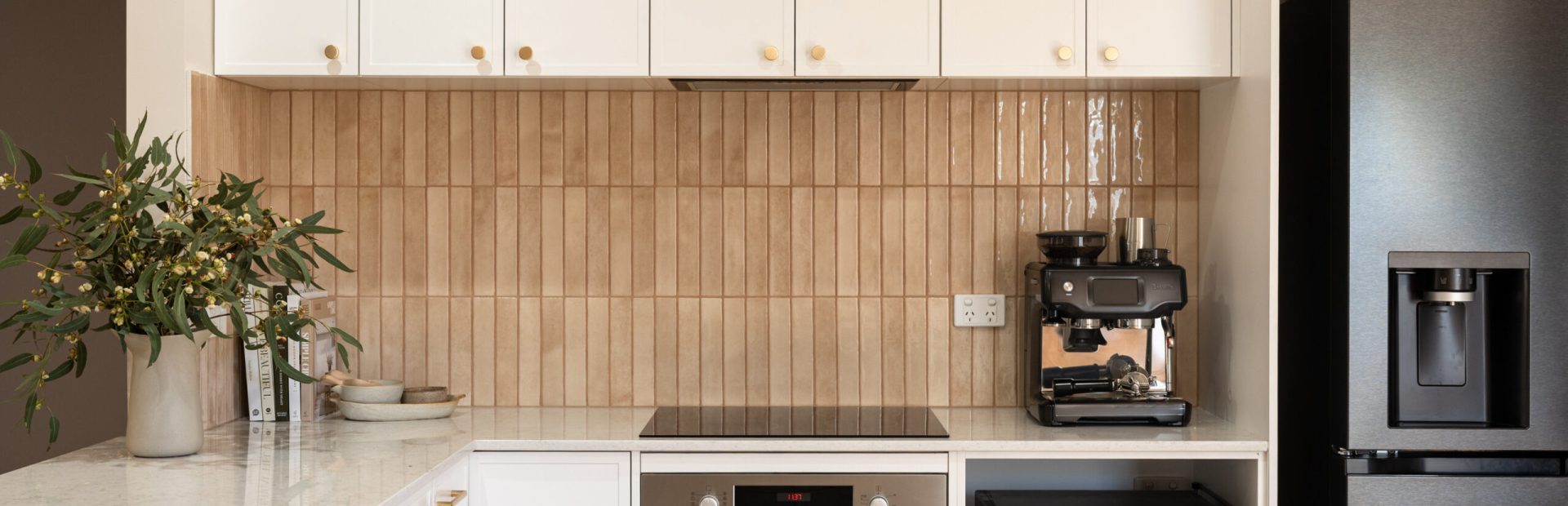 U shape kitchen displaying hampton style white kitchen cabinetry with brass handles and pink tiled splashback with coffee machine on stone benchtops and black oven and microwave
