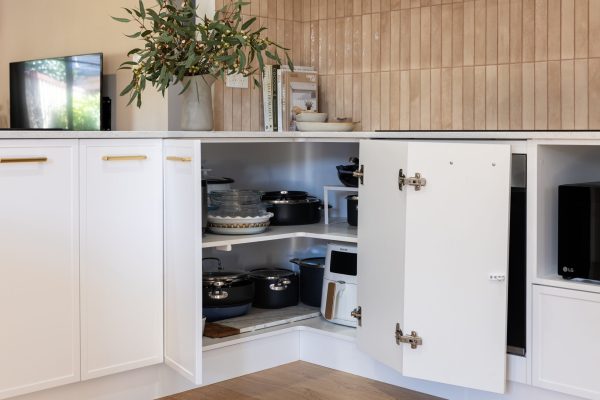 Corner cupboard in u shape kitchen displaying pots and pans with brass handles and plant and cookbooks styling on stone benchtops