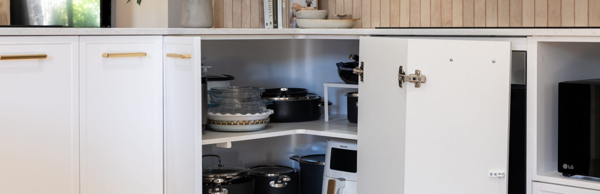 Corner cupboard in u shape kitchen displaying pots and pans with brass handles and plant and cookbooks styling on stone benchtops