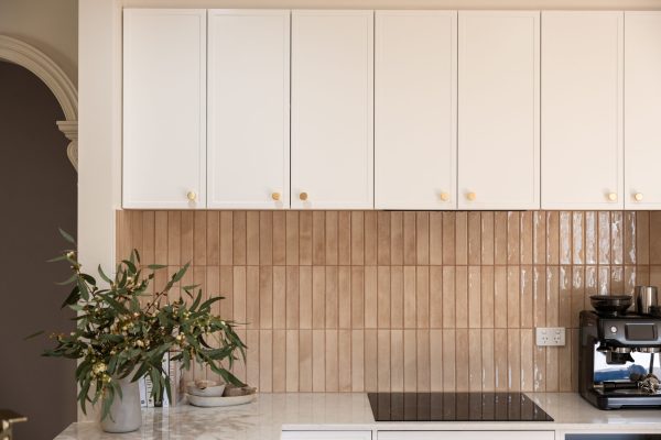 White colour hampton style kitchen cabinetry overheads with brass knob handles and cream tiled splashback with black cooktop and plant on benchtop