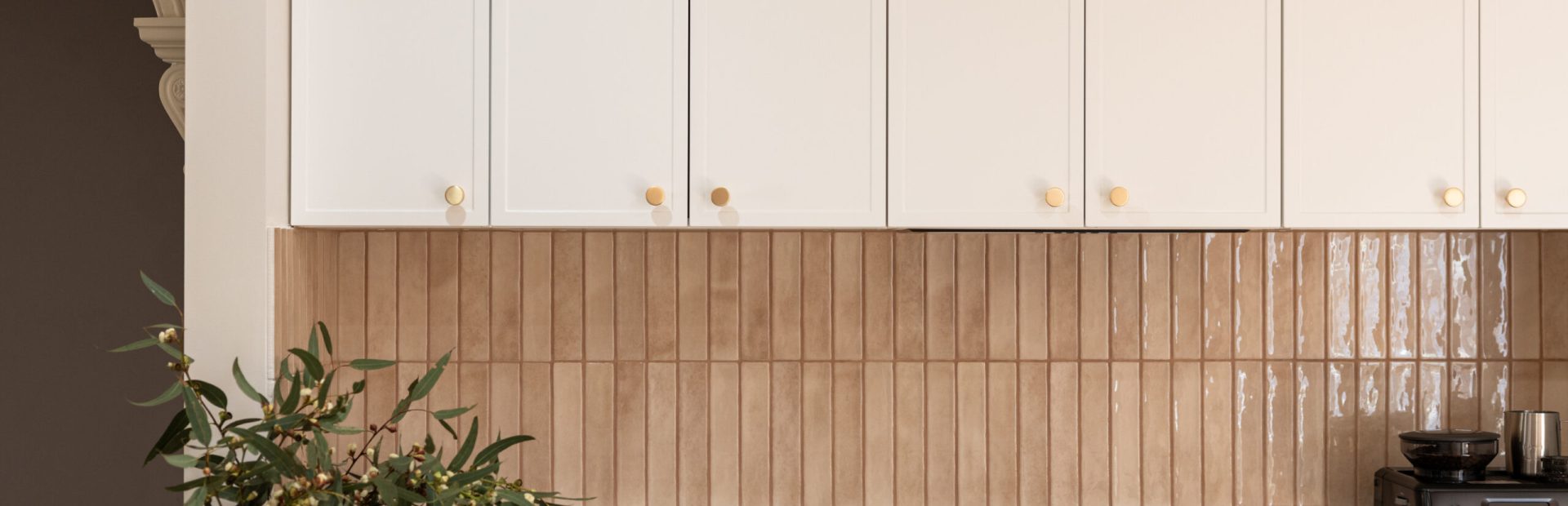 White colour hampton style kitchen cabinetry overheads with brass knob handles and cream tiled splashback with black cooktop and plant on benchtop