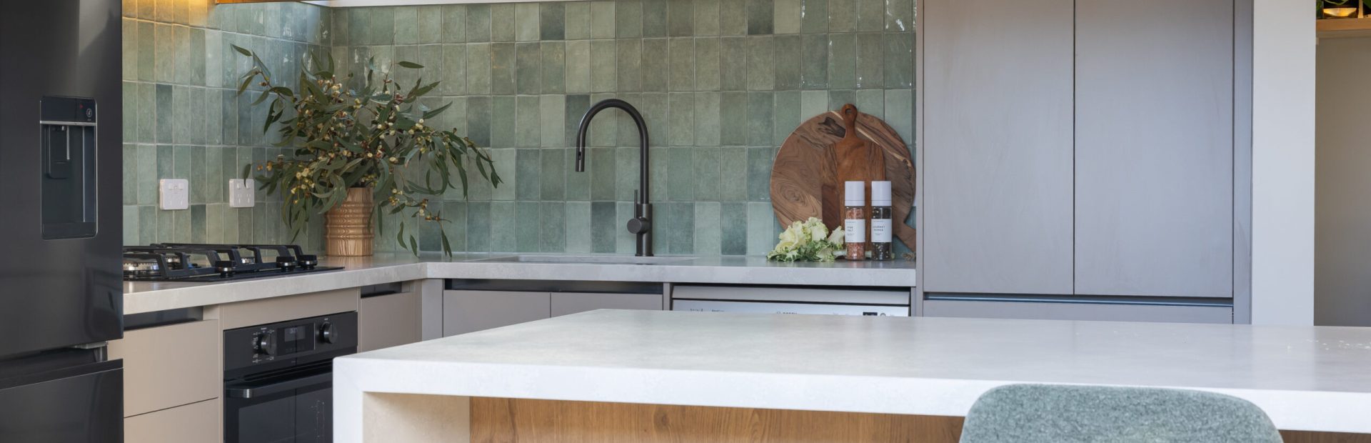 Kitchen displaying pantry in beige coloured cabinetry with green tiled splashback and chopping board and plants decorated on benchtops