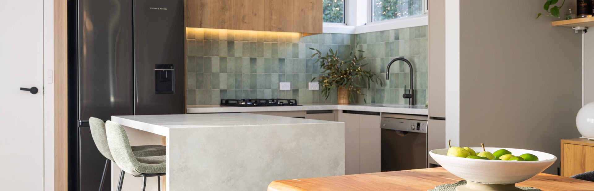 U shape kitchen with black friedge and sink and tap and oak overhead cabinetry with green tiled splashback and standalone island bench in stone