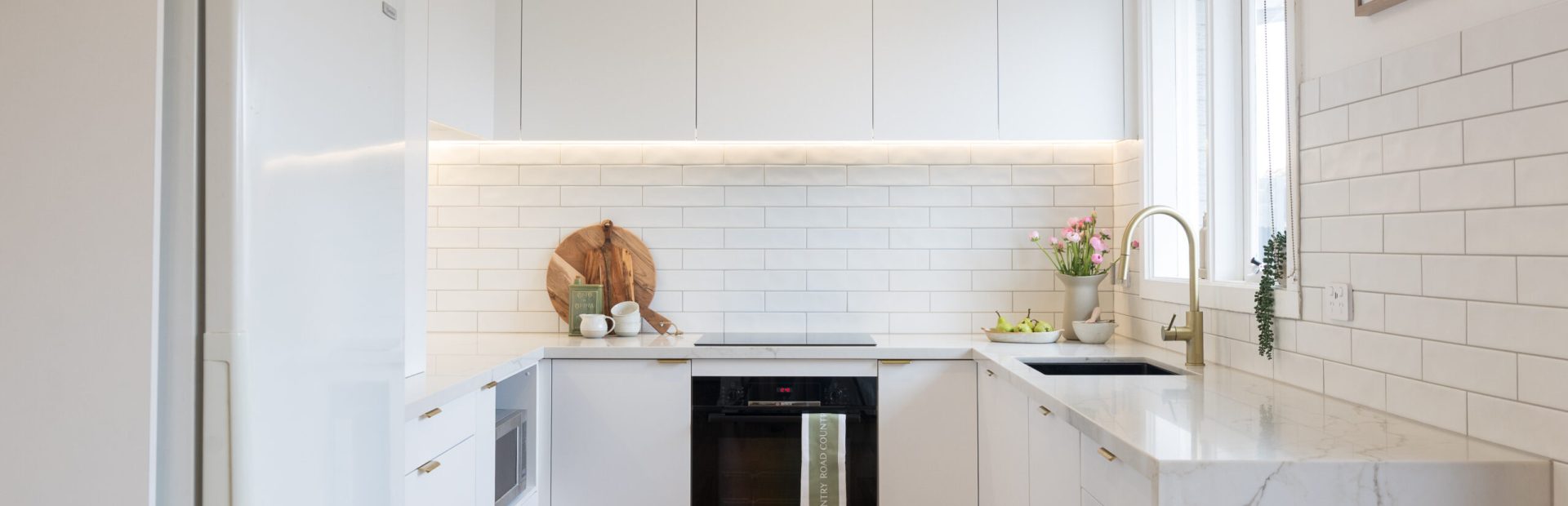 U shaped white kitchen with white and vein stone benchtop with wooden chopping boards and plants on kitchen benchtop for styling