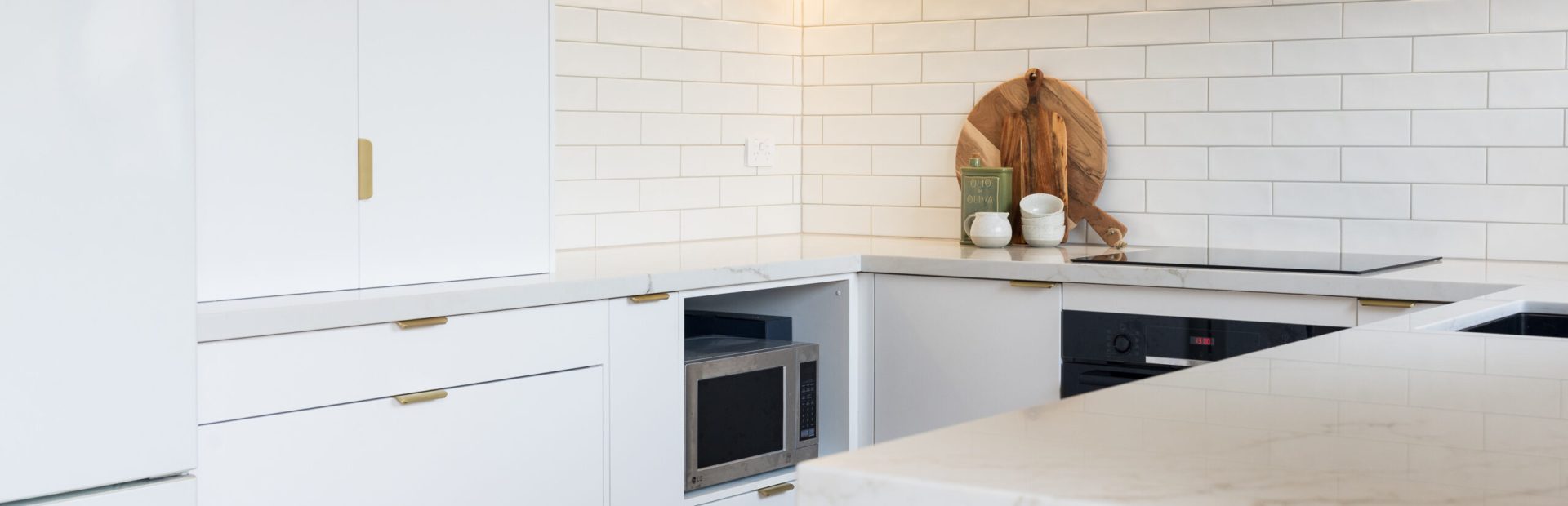 White kitchen displaying a microwave cabinet and appliance cabinet with brass handles and kitchen styling items on benchtop