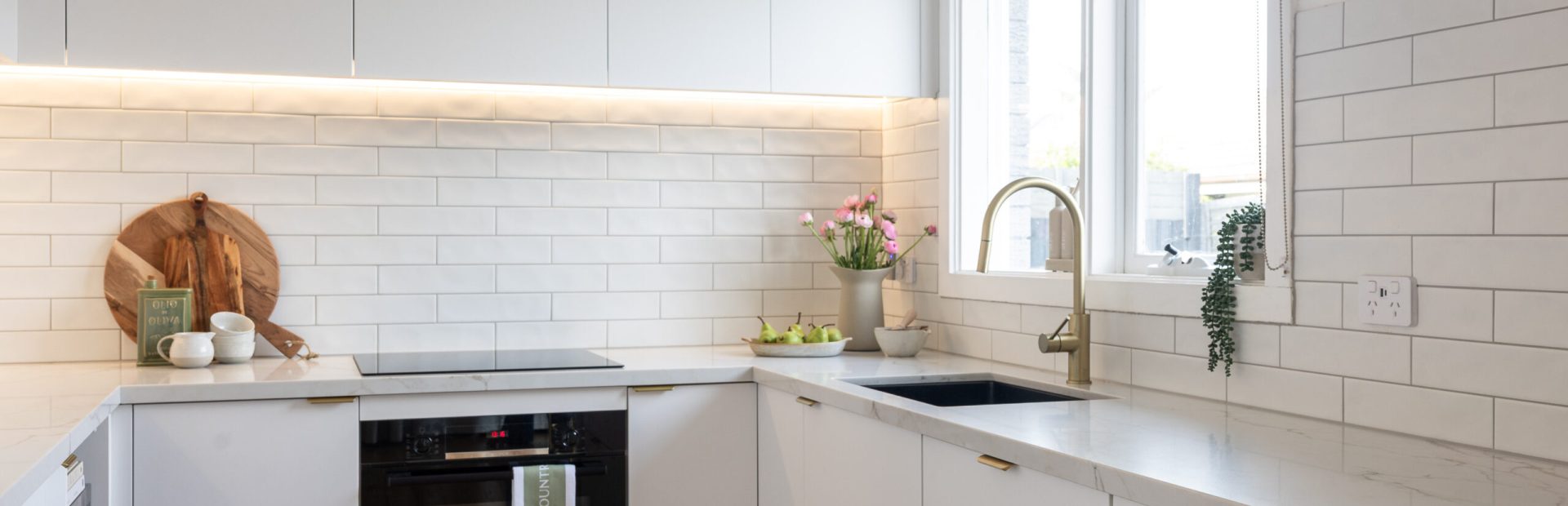 Kitchen displaying white cabinetry with stone veined benchtops and brass handles and tapware with plants for styling