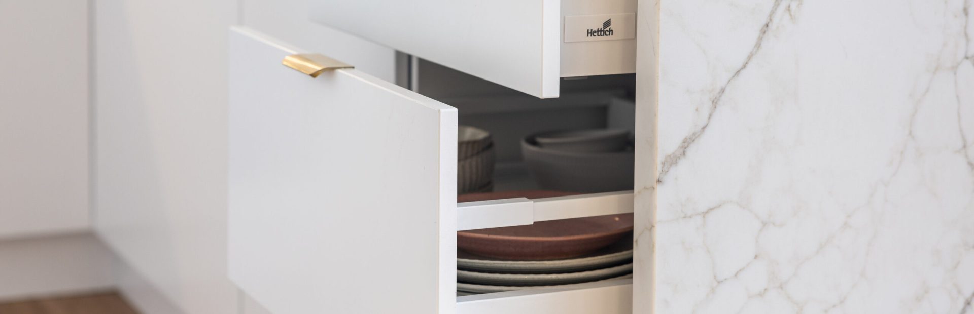White kitchen displaying two pull out drawer sizes with brass handles and plates and bowls