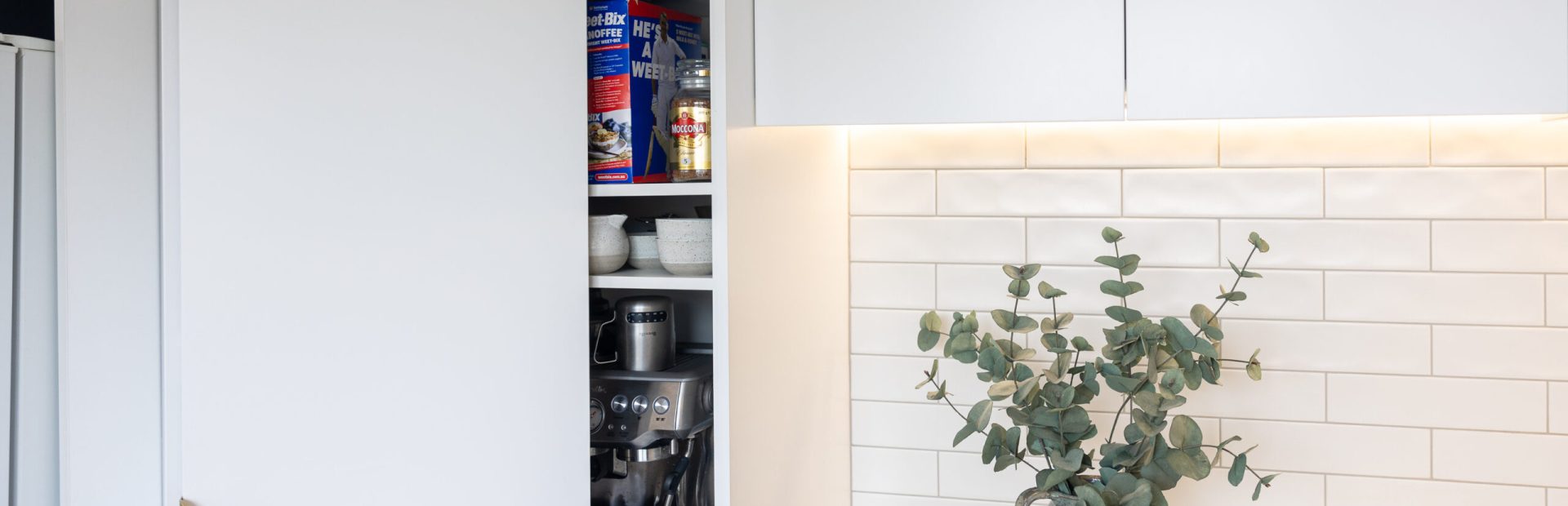 White kitchen pantry cabinetry with styling books and grey jug with green folliage on stone benchtop