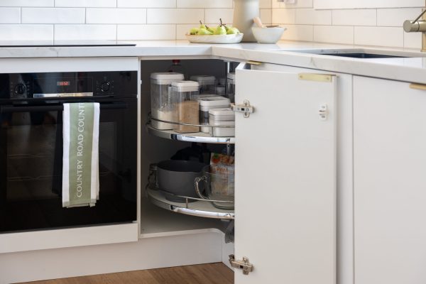 Corner cabinet in a white kitchen displaying a lazy susan with pantry items and pots next to a Country Road tea towel