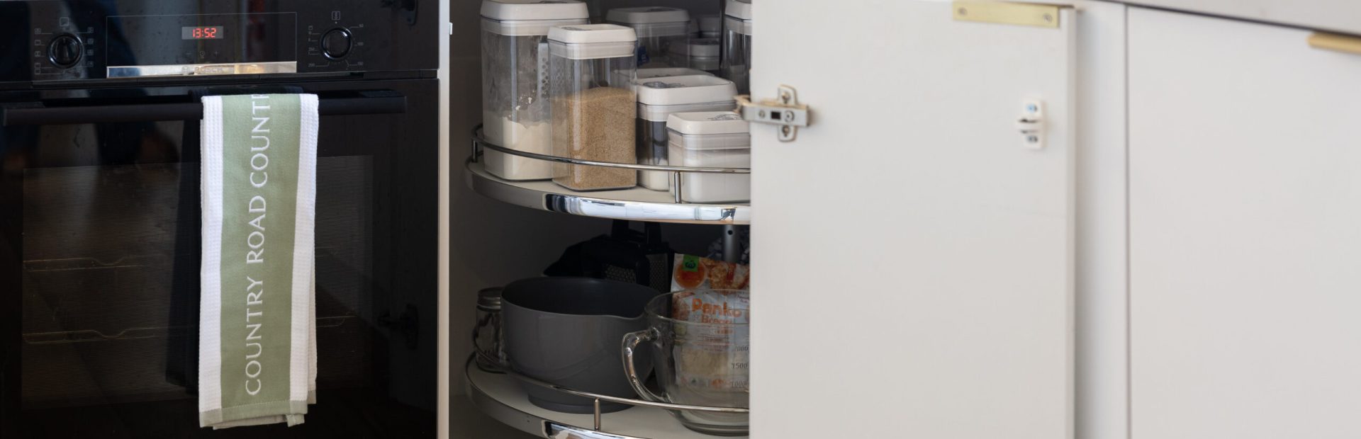 Corner cabinet in a white kitchen displaying a lazy susan with pantry items and pots next to a Country Road tea towel