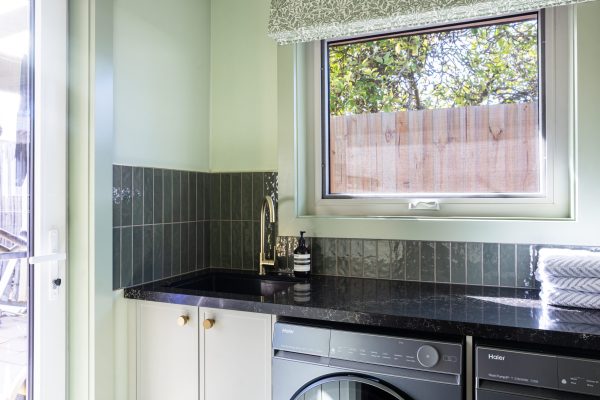 Galley laundry featuring green walls, tiles and curtains with brass tap and handles, also featuring black benchtop and parchment colour hampton cabinetry