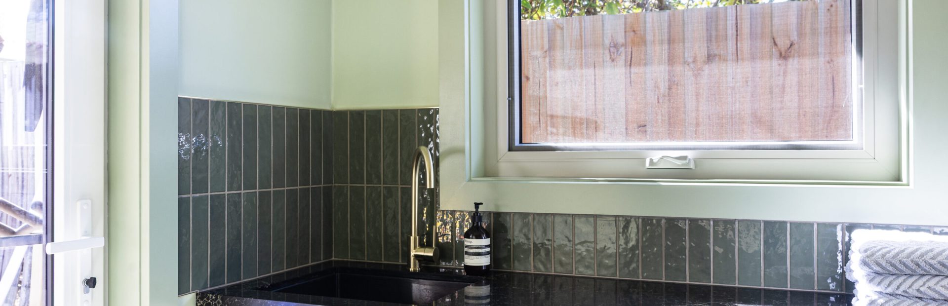 Galley laundry featuring green walls, tiles and curtains with brass tap and handles, also featuring black benchtop and parchment colour hampton cabinetry