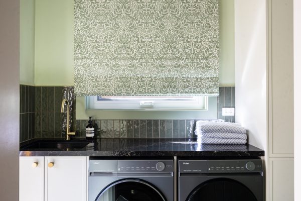 Laundry featuring hampton style cabinetry with washer and dryer side by side with black benchtops, green tiled splashback and brass taps and handles