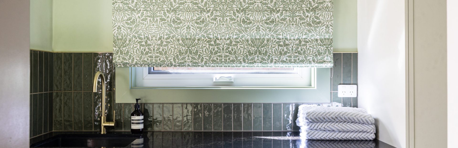 Laundry featuring hampton style cabinetry with washer and dryer side by side with black benchtops, green tiled splashback and brass taps and handles