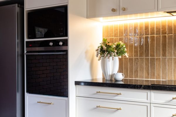 Hampton kitchen cabinetry in parchment colour with brass handles, displaying wall oven tower with black ovens and next to black fridge set against brown tiled splashback