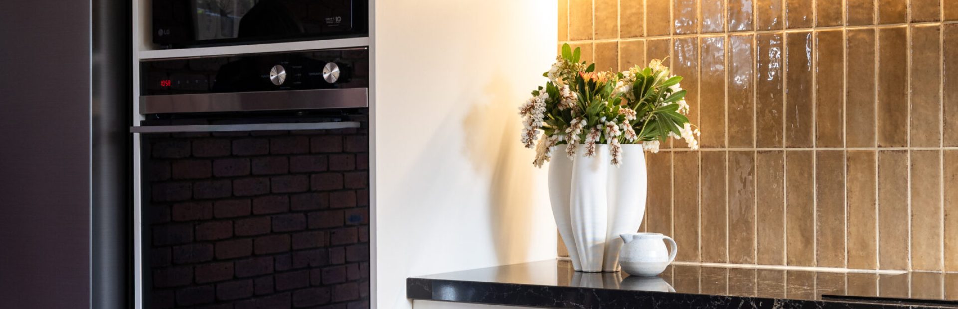 Hampton kitchen cabinetry in parchment colour with brass handles, displaying wall oven tower with black ovens and next to black fridge set against brown tiled splashback
