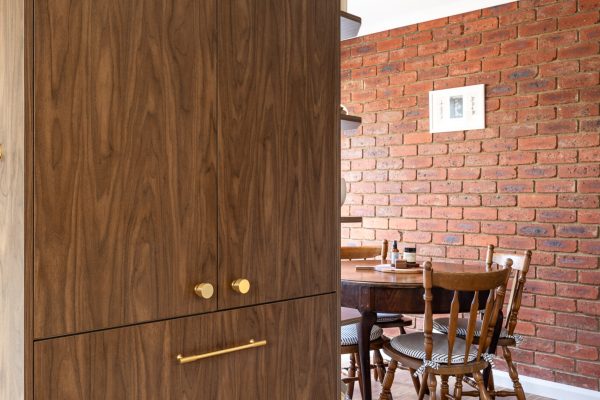 Kitchen displaying sepia walnut colour cabinetry in pantry with brass handles and timber round dining table with brick feature wall