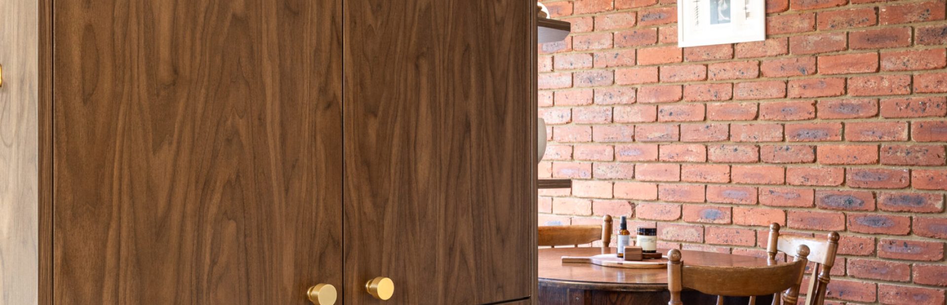 Kitchen displaying sepia walnut colour cabinetry in pantry with brass handles and timber round dining table with brick feature wall