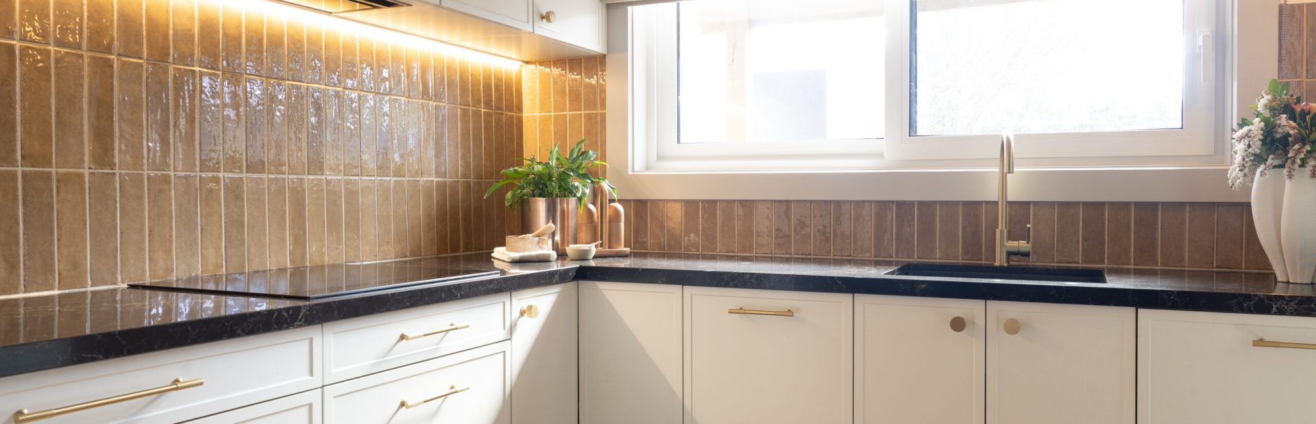 Hampton style kitchen with parchment cabinetry and brass handles with black benchtops displaying brass pot plant and styling items on bench