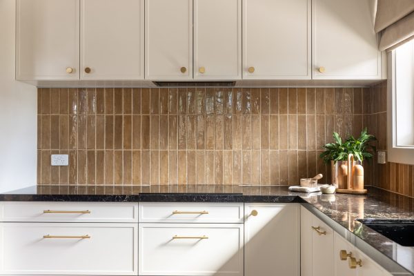 Parchment colour kitchen cabinetry with brown tiled splashback, brass handles and black benchtops with bowls and pot plant styling the kitchen