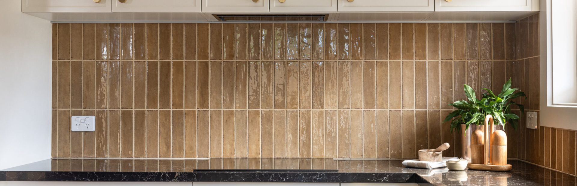 Parchment colour kitchen cabinetry with brown tiled splashback, brass handles and black benchtops with bowls and pot plant styling the kitchen
