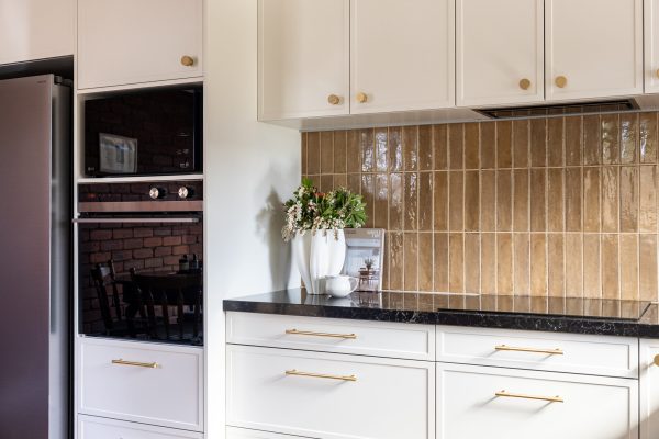 Brown tiled splashback displaying hampton style cabinetry in parchment colour with brass handles, black benchtops and appliances styled with vase of flowers, bowl and cookbook
