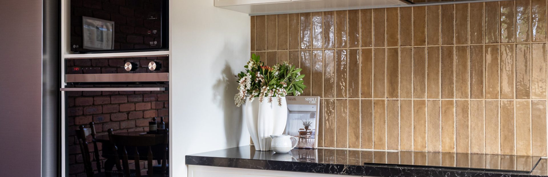 Brown tiled splashback displaying hampton style cabinetry in parchment colour with brass handles, black benchtops and appliances styled with vase of flowers, bowl and cookbook