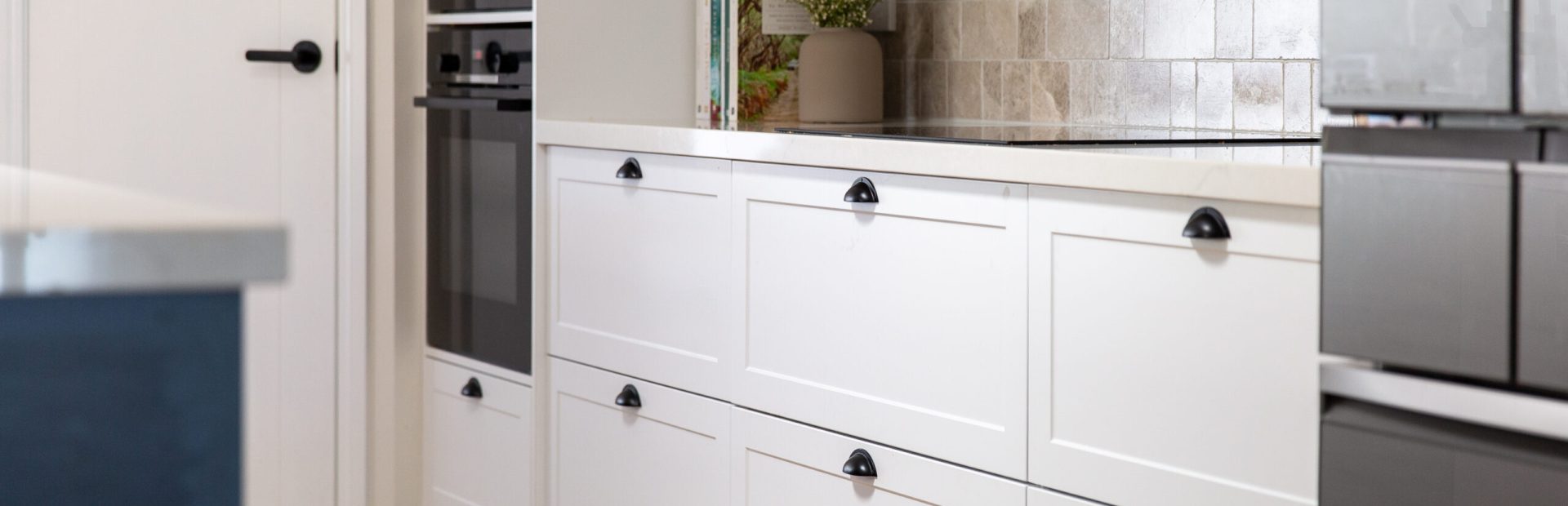 Long row of white shaker style cabinetry with black handles and black wall ovens