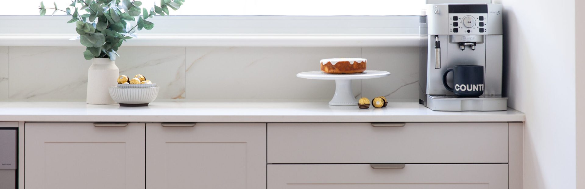 Light grey Shaker style kitchen cabinetry featuring doors and drawers with chrome handles and coffee machine with cake on stone benchtop