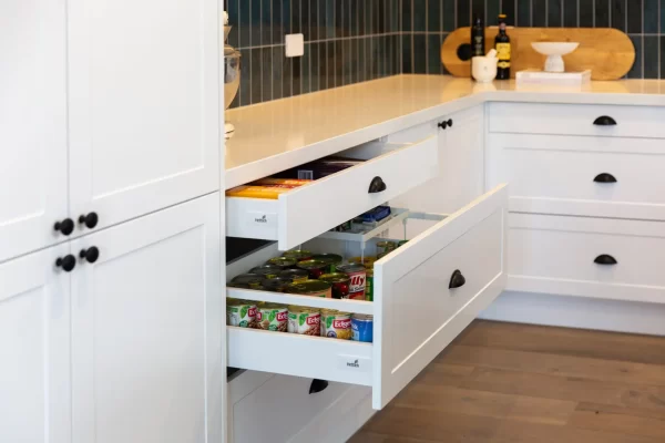 White shaker style kitchen drawer cabinets featuring tins of canned food and white stone benchtops with green tiled splashback
